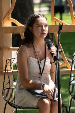 A teen girl with long brown hair and a white sundress speaks into a microphone on an outdoor stage.