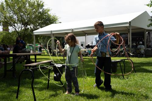 A young brother and sister stand outside in the grass on a sunny day with a white tent in the background behind them. They hold rope lassos and prepare to lasso a small model of a steer.