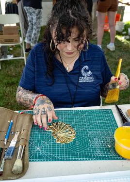 A woman with gold hoop earrings and tattooed arms holds a paint brush in one hand and positions a golden fan-shaped ornament with the other. Beside her are more paintbrushes and a tub of yellow paint.