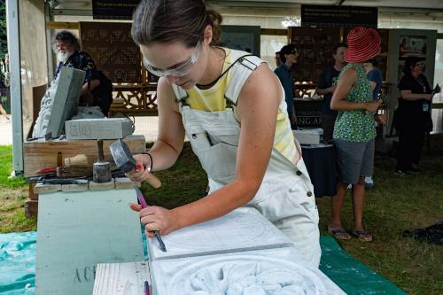 A young woman in white overalls and safety glasses uses a chisel in one hand and hammer in the other to carve a circular pattern in a block of white stone. Behind her, festival visitors explore the displays under a tent.