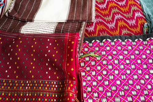 Four woven textile arranged on a table: brown and white stripes; red with yellow, white, and green wavy stripes; red with a black diamond pattern and yellow, white, and green accents; and pink checkerboard pattern on black with yellow diagonal stripes and
