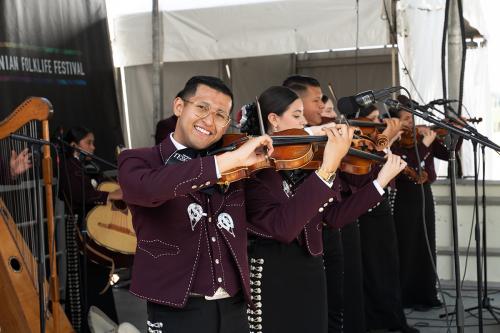 Several musicians play string instruments on an outdoor stage, wearing matching maroon jackets and black pants or skirts. The musician in front smiles broadly at the camera.