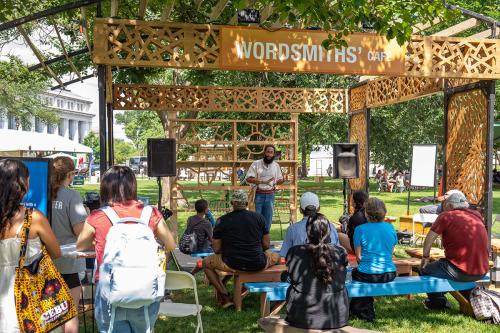A wooden lattice structure in the grass among trees, with a sign labeling it the Wordsmiths’ Cafe. Below the lattice, a man stands and performs in front of a small seated audience in dappled sunlight.
