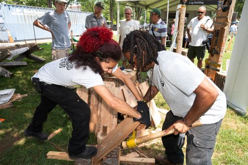Two young men stand facing each other, hunched over working to split a wooden log. Their faces show the effort and the heat of the day. Behind them, a group of individuals observes, including a person in a National Park Service uniform. 