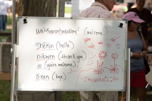 Words and translations on a whiteboard outdoors: wa'tkawnonhweraton'! (welcome!) she:kon (hello). nia:wen (thank you). io (you're welcome). o:nen (bye). Plus a drawing of flowers with roots in the ground under stars, hearts, and smiley faces.
