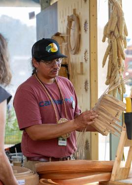 A person holds up a woven basket in progress. Behind him, ears of corn are strung together, hanging down to dry.