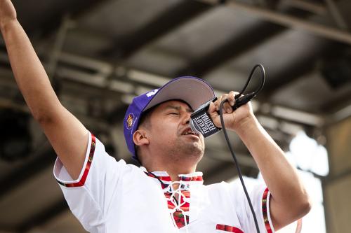 A person wearing a purple hat and a white shirt with red details closes his eyes and sings into a microphone onstage.