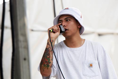 A person wearing a gray Carrhart t-shirt and a white bucket hat speaks into a wired microphone. A colorful tattoo is visible wrapping around his forearm.