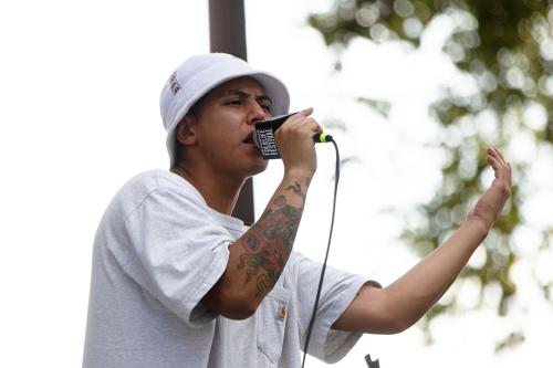 A person wearing a white bucket hat and a gray t-shirt sings into a microphone. A colorful tattoo is visible on his forearm.