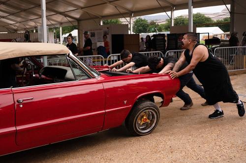 Three people push the front end of a red classic car down a dirt path in front of a large tent.