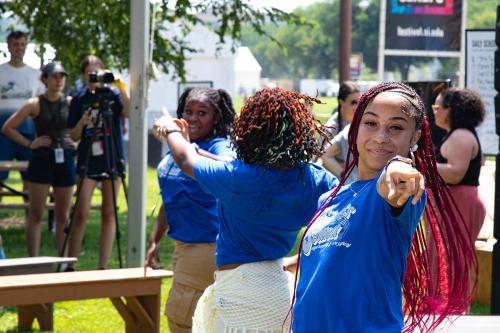 Three dancers in matching blue T-shirts dance in the grass. The dancer in front looks straight at and points to the camera with a smile on her face.