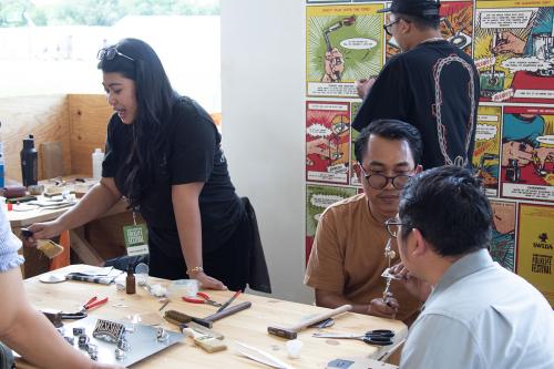 Around a work table set with metalworking tools, people chat.