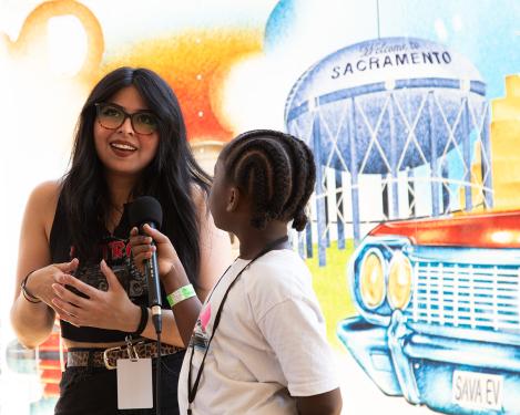 A young girl with curved cornrows holds a microphone up to a woman with long dark hair and glasses. Behind them is a mural with the front of classic red car and a water tower that says Welcome to Sacramento.