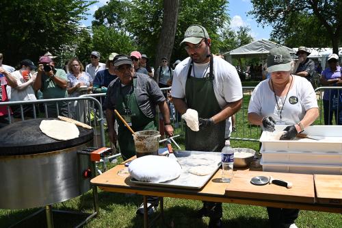 Three people work on at an outdoor cooking station, handling circles of dough that are then placed on a convex oven. A crowd of people watch from behind low gates.