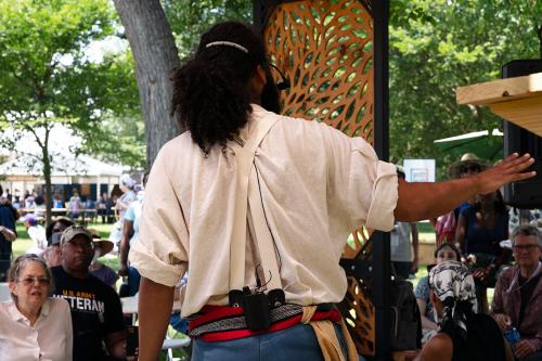 From  behind, the man performs and gestures outward toward a seated audience. Although he is dressed in old-timey clothes, he has a wireless microphone device clipped to his belt.