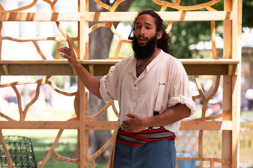 The man performs on the wooden stage, lifting one hand with index finger pointed upward.