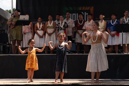 Three young girls stand side by side. From left to right, each is taller than the last. They wear Hawaiian leis and are demonstrating motions of the hula to an audience. Behind them on the mainstage, other members of their group, both old and young, stand