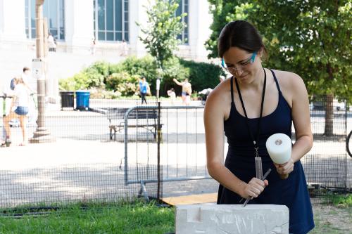 A young woman wearing a black tank top and safety glasses uses a hammer and chisel to carve into a stone block outdoors.