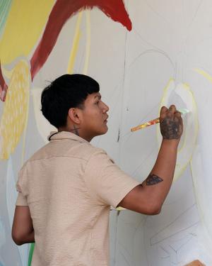 A young man with tattoos on her hand, forearm, and neck uses a paintbrush to outline vanilla flowers on a large outdoor mural.