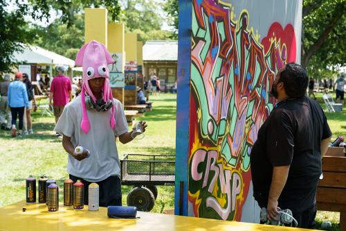Two people stand on either side of a free-standing wall covered in colorful graffiti art. One holds a respirator, and the other holds a plastic cup and a can of spray paint and wears a silly pink squid hat.