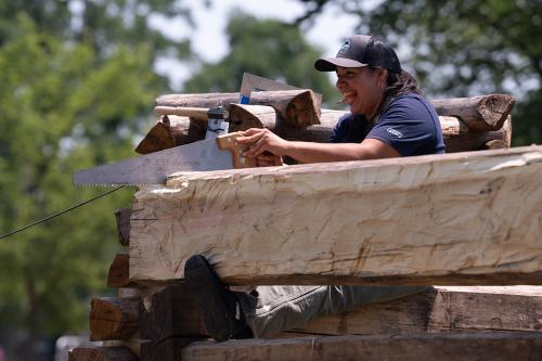 A young woman balances herself between two wooden logs. She uses a Disston brand saw to saw a notch into the top of one of the logs. She holds a pencil in her teeth and has a big smile on her face. She wears a baseball cap that has a logo reading Stewards