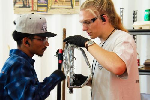 Two young men work on either side of a bare bike frame set on a stand. They war latex gloves and safety glasses.
