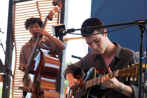 In the foreground, a young person who is seated on a stage looks down at the Eastman guitar he is playing. In the background behind him, another young bandmate plays a stand-up bass and looks off into the distance.