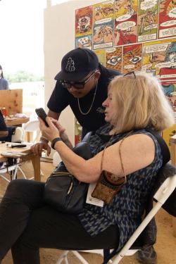 A blonde woman is seated at a wooden table under a white tent, and a man wearing a black baseball cap looks at something she is pointing at one her smartphone.