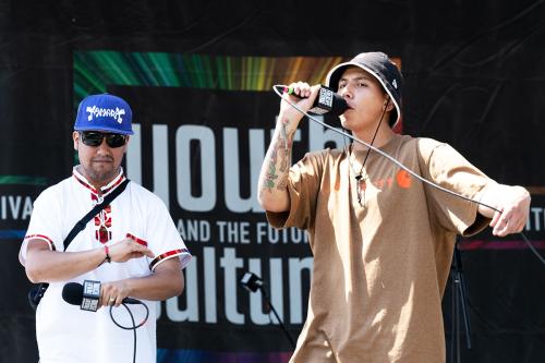 Two people stand on a stage holding wired microphones. One wears a white shirt and a blue baseball cap that reads AYMARA, holding his microphone at his chest. The other wears a brown t-shirt and an orange bucket hat and raps into the microphone.