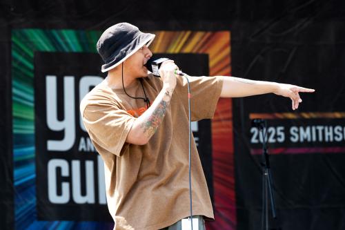 A person in a gray bucket hat and brown t-shirt is singing onstage into a microphone, pointing with his arm extended to his left.