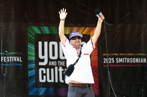 A person wearing a white shirt with red details and a purple baseball cap put his both his hands up in the air while holding onto a wired microphone onstage.