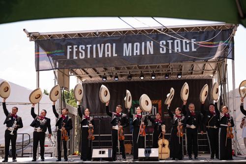Eleven musicians in matching black suits and maroon ties wave their wide brimmed hats from on stage, under a banner reading Festival Main Stage.