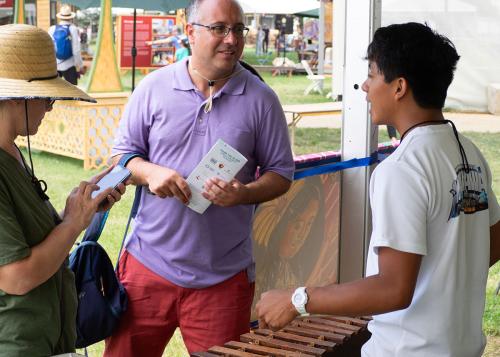A young man stands in front of a wooden marimba, talking with two festival visitors.
