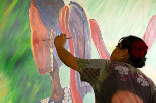 From behind, a woman paints the long dark hair of a woman in the mural. The two women face the same direction.