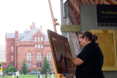 A woman with long brown hair in a low bun and a black polo shirt works on an art piece on an easel. In the background is one wing of the red-brick Smithsonian Castle.