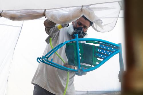 A man wearing a respirator uses a pneumatic spray gun to paint the frame of a bike shiny blue. Adornments in the frame of the bike form the stars and stripes of the American flag.