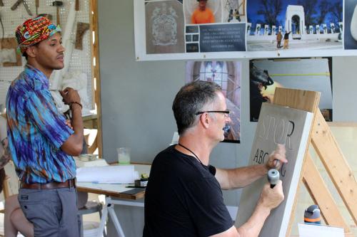 A man sits at a makeshift easel with a chisel in one hand and hammer in the other, carving into the letter R on a gray stone tablet. Another person stands behind him to watch.