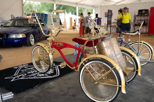 A red lowrider tricycle with golden handlebars, fenders, and spokes and a cyclindrical tank attached below the back of the saddle.The word Olathe is painted in gold and black on the side of the frame.