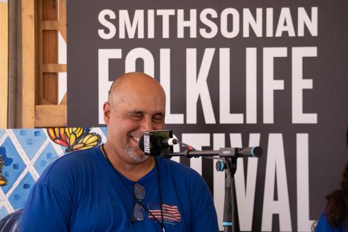 A man wearing a bright blue t-shirt sits behind a microphone onstage. His eyes are squeezed tight and he smiles while laughing. Behind him, the Smithsonian Folklife Festival logo is visible.