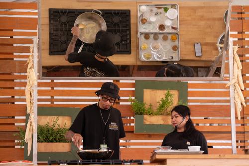 A man and a woman, both wearing black t-shirts, stand onstage behind a counter with a gas stove on top. The man, who wears a black bucket hat, stirs something in a metal pan, while the woman looks on. All of this is reflected to an audience in an overhead