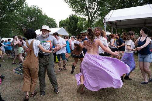 A large group of people, divided into pairs, swing their partners around on a grass dance floor. On the left of the photo, two older people dance together, while on the right, two young people, one in a flowing lavender dress, spin each other around.