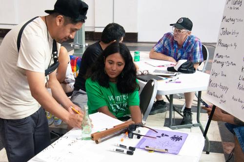 A person in a cream colored t-shirt wearing a black baseball cap backwards holds a pencil and gestures towards a piece of paper on a table. A person in a green t-shirt that says VOLUNTEER listens intently and looks at the paper.