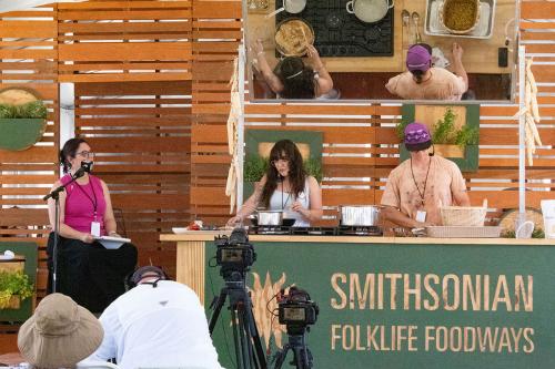 Two people cook on an outdoor stage, while a third person speaks into a microphone and watches. On the stove is a pot of corn mush. Also on the counter is a woven basket.