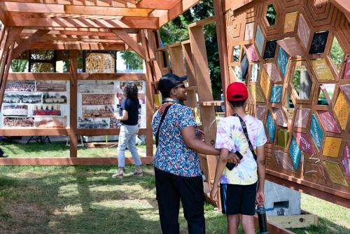 An adult woman, who's grinning and wearing a floral blouse, and adolescent boy, who's wearing a red baseball cap and tie-dye t-shirt, look at a wooden wall of colorful tiles displaying various terms in slang through generations.