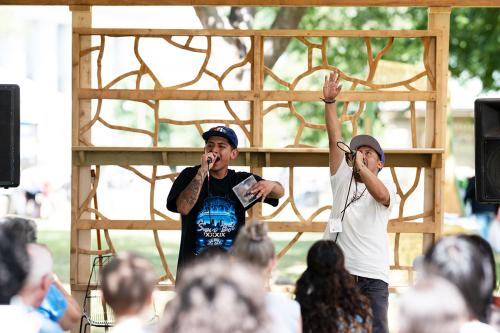 Two people stand outdoors in front of a wooden structure while singing into wired microphones. One person looks up towards the sky and raises his hand up to the sky. The other holds onto a CD in his hand.