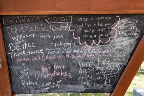 A chalkboard wall displays advice written by festival guests to their younger selves; Don't touch your eyebrows and Care a lot + be silly are two examples.