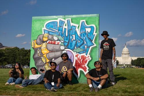 Six people pose around a freestanding mural in the grass, with the U.S. Capitol Building in the distance. The mural includes the word SWEDA in graffiti script and a hand adorned with many gold rings. The rings each contain a word, spelling out Folk Life F