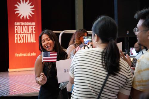 A young woman holds a small American flag and sticks her tongue out while closing her eyes, posing for a photo taken on a smartphone. Behind her onstage, the Smithsonian Folklife Festival logo is visible.