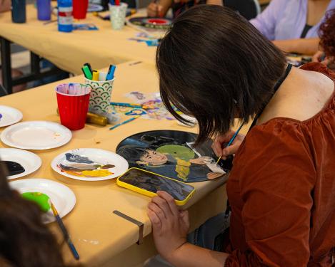 A young woman is seated at a table, facing away from the camera. She leans over her work, as she carefully paints on an old vinyl record. Next to the record on the table is her smartphone, which displays the image she is recreating in paint on the record—