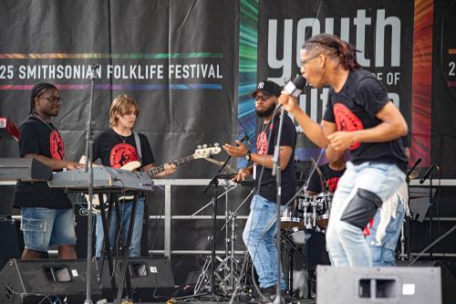 On stage, four teen musicians play keyboard, bass, and drums, and sing. An older man appears to play air bass.